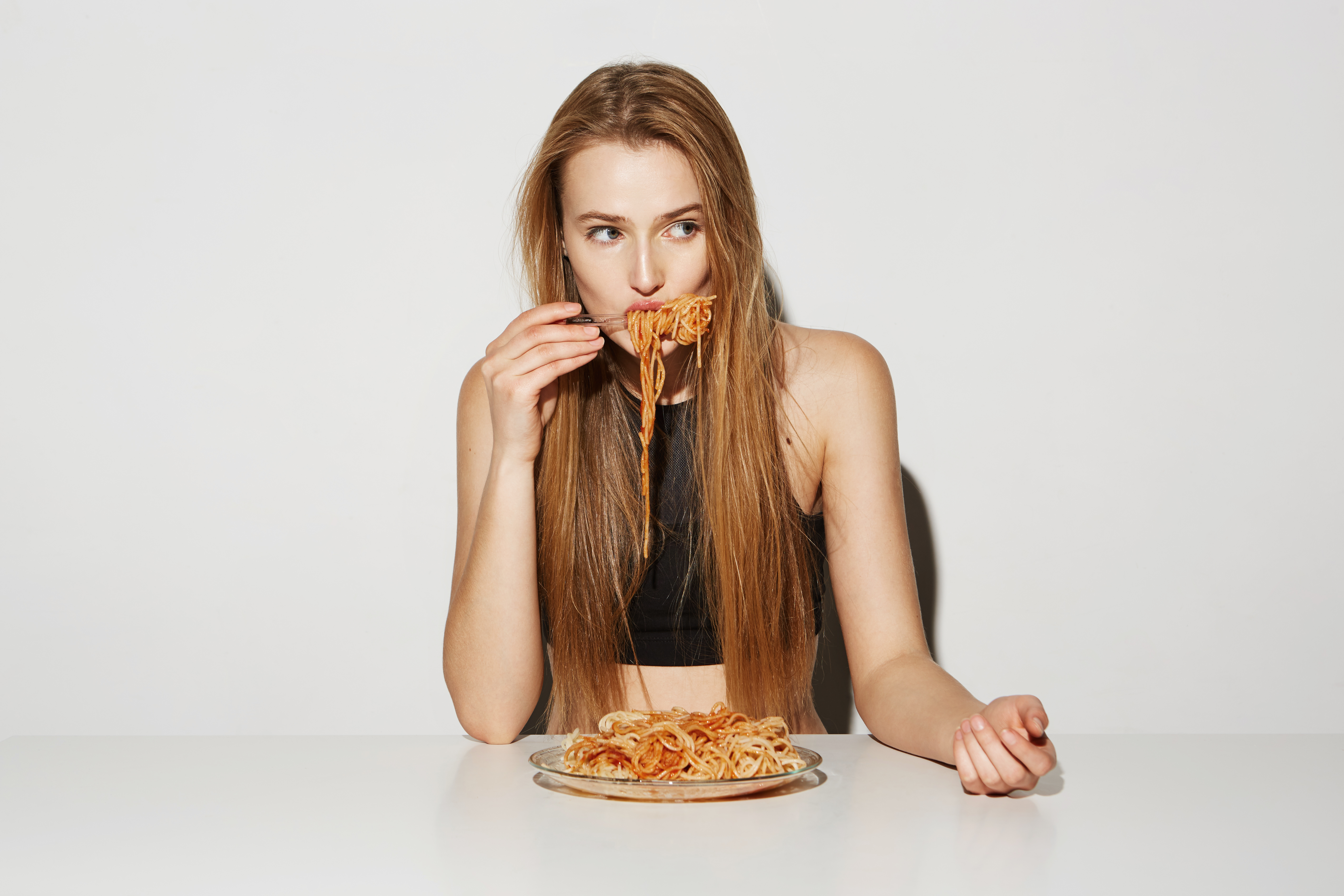 Close up portrait of sexy blonde girl with long hair sitting at table, eating spaghetti, looking aside with relaxed and flirty expression.